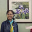 Piehong E. stands in front of her art which depicts flowers in front of her painting which depicts purple iris flowers